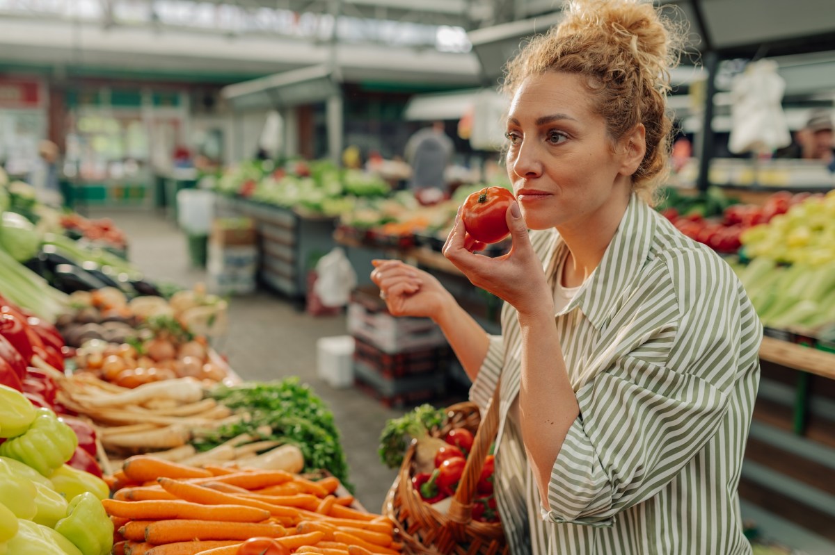 Vegane Mythen: eine Frau steht am GemÃ¼sestand und hÃ¤lt eine Tomate in der einen und einen Einkaufskorb in der anderen Hand.