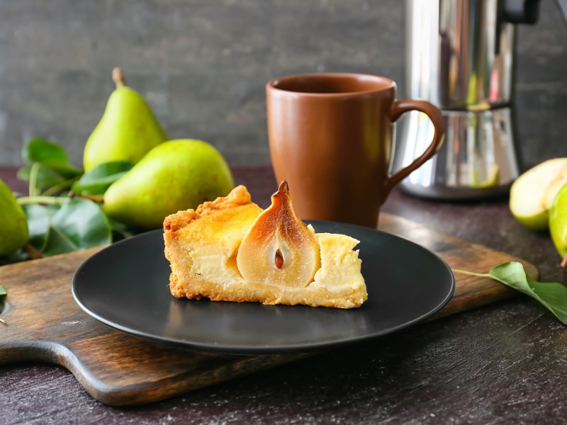 Ein Stück Birnen-Schmand-Kuchen mit einer halben Birne auf einem dunklen Teller.