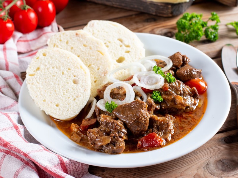 Ein Teller mit Gulasch und bÃ¶hmischen KnÃ¶deln, daneben Besteck und ein kariertes Geschirrtuch.