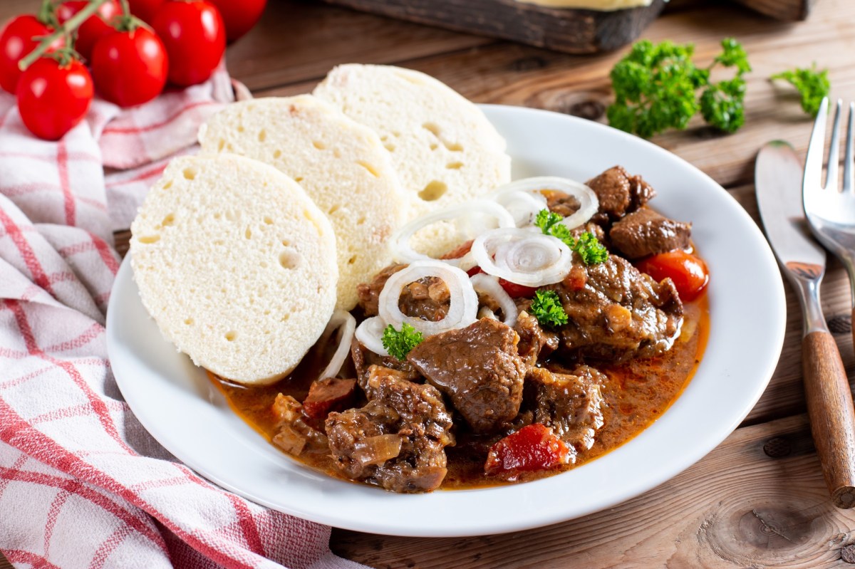 Ein Teller mit Gulasch und bÃ¶hmischen KnÃ¶deln, daneben Besteck und ein kariertes Geschirrtuch.