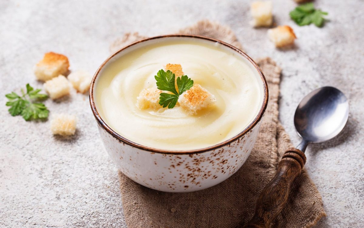 Eine SchÃ¼ssel Apfel-Blumenkohlsuppe mit CroÃ»tons, drumherum weitere CroÃ»tons und ein LÃ¶ffel.