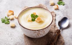 Eine Schüssel Apfel-Blumenkohlsuppe mit Croûtons, drumherum weitere Croûtons und ein Löffel.