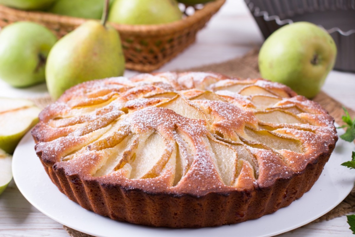 Ein Birnenkuchen mit Puderzucker auf einem Teller. Im Hintergrund liegen frische Birnen.