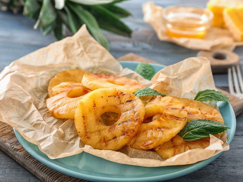 Ein hellblauer Teller ausgelegt mit Backpapier auf dem Ananasringe aus dem Airfryer liegen. Garniert sind die Ananasringe mit frischer Minze. Im Hintergrund steht eine kleine Schale mit Honig und die BlÃ¤tter einer Ananas sind zu sehen.