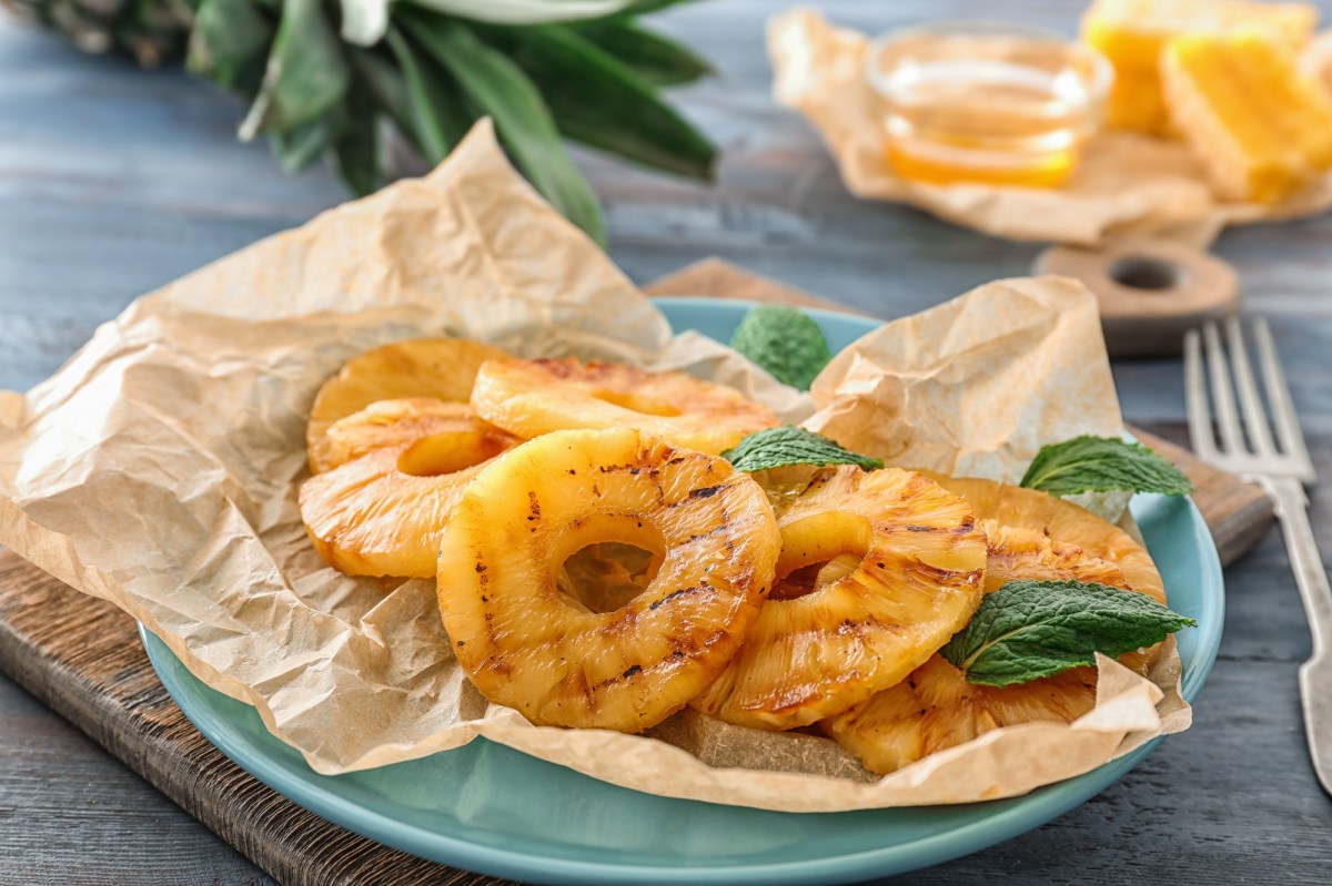 Ein hellblauer Teller ausgelegt mit Backpapier auf dem Ananasringe aus dem Airfryer liegen. Garniert sind die Ananasringe mit frischer Minze. Im Hintergrund steht eine kleine Schale mit Honig und die BlÃ¤tter einer Ananas sind zu sehen.