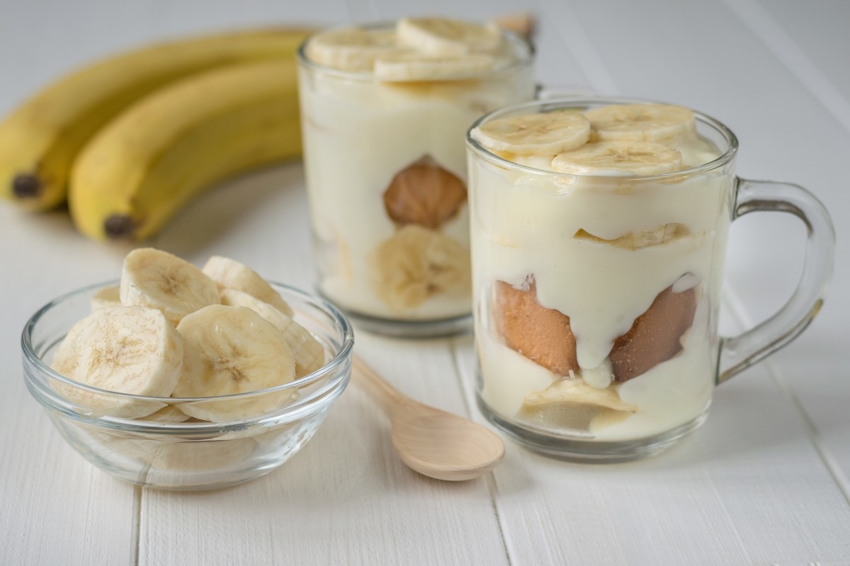 Zwei DessertglÃ¤ser mit Banana-Pudding. Daneben steht ein kleines SchÃ¤lchen mit Bananenscheiben. AuÃŸerdem liegen zwei Bananen und ein HolzlÃ¶ffel dabei.