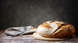 Ein Brot aus drei Zutaten liegt auf einem runden Brett auf einem schlichten Holztisch. Links daneben liegt ein Tuch sowie ein Brotmeser.