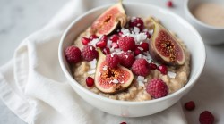 Eine Schüssel mit Chai-Porridge mit Granatapfelkernen, Feigen, Himbeeren und Kokosflocken. Die Schüssel steht auf einem weißen Küchentuch. Es liegen einzelne Granatapfelkerne und Himbeeren verstreut. In der linken oberen Bildecke kann man verschwommen eine kleine Schale mit Zimtzucker oder Milchkaffee erkennen.