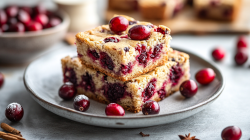 ein paar Cranberry-Blondies auf einem Teller, drumherum frische Cranberrys und einige Zimtstangen. Im Hintergrund eine Schale mit mehr Cranberrys und ein Tablett mit weiteren Blondies.