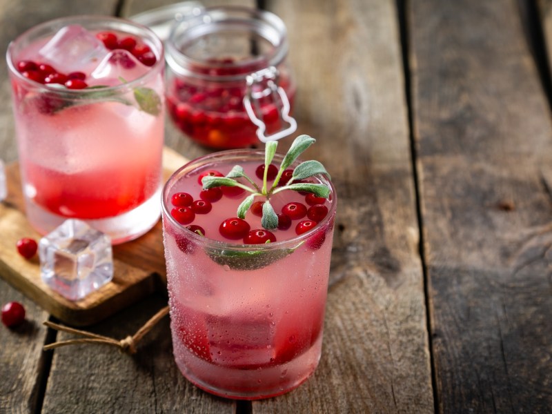 Zwei GlÃ¤ser Cranberry-Salbei-Mocktail. Im Hintergrund steht ei Einmachglas mit Cranberrys darin.