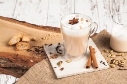 Eine Glastasse mit Dirty Chai Latte einem weißen Untersetzer. Der Untersetzer mit der Glastasse steht auf einem Stück Holz, das mit Sackleinen bedeckt ist. Außerdem liegen Cantuccini rum, Zimtstangen, Nelken und andere Chai-Gewürze.