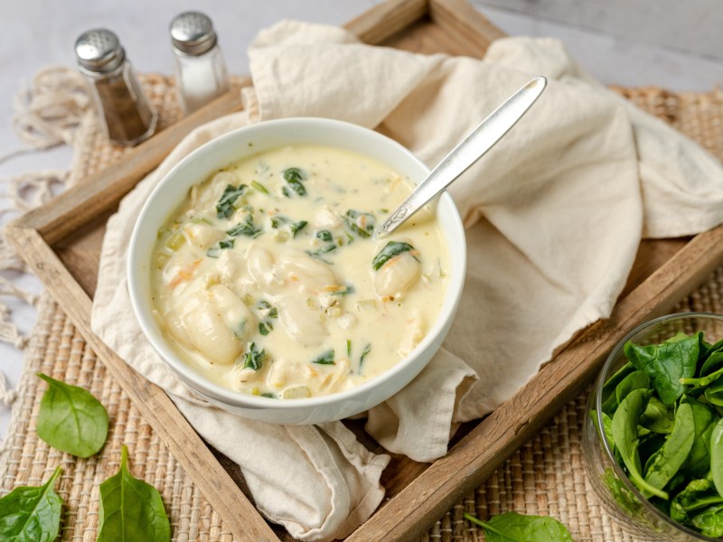 Eine SchÃ¼ssel voll cremiger Gnocchi-Suppe mit Artischocken und Spinat serviert auf einem Holzbrett ausgelegt mit einer beigen Serviette. Daneben ist ein Salz- und Pfefferstreuer zu sehen. AuÃŸerdem steht eine kleine Schale mit SpinatblÃ¤ttern daneben.