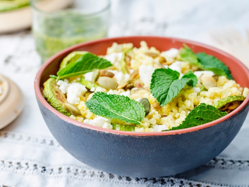 Hirsesalat mit Ziegenkäse in einer . Angerichtet mit frischen Minzblättern in einer schwarz-roten Schüssel auf einem weißen Tuch.