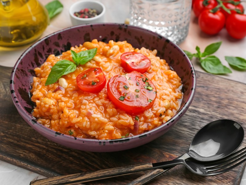 Italienischer Tomatenreis mit Bohnen in einer SchÃ¼ssel, garniert mit Tomatenscheiben und Basilikum. Drumherum einige Zutaten.