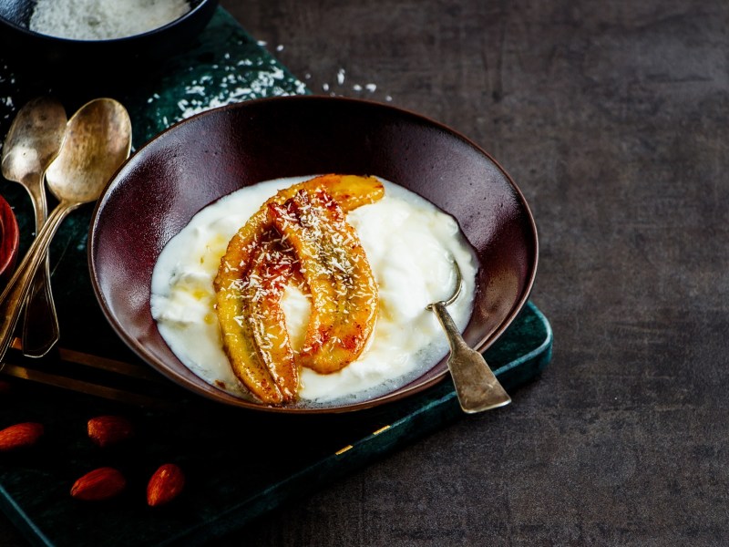 Eine SchÃ¼ssel voll Joghurt mit karamellisierten Bananen. Garniert mit Kokosraspel. Daneben liegen LÃ¶ffel und ein paar Gojibeeren.