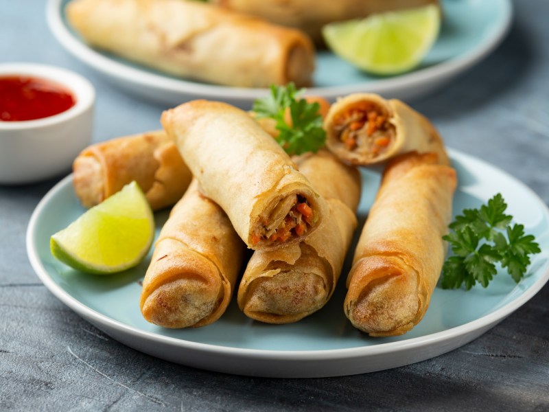 Kimchi-Frühlingsrollen liegen auf einem hellblauen Teller. Sie sind mit einem Limettenstück und frischer Petersilie garniert. Im Hintergrund steht eine Schale mit Soße, sowie ein weiterer Teller Frühlingsrollen.