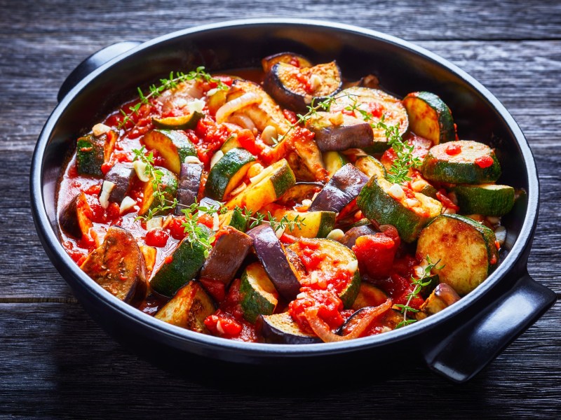 Ein GemÃ¼seeintopf mit Auberginen, Zucchini, Tomaten, Knoblauch und Zwiebeln, serviert in einer schwarzen Keramikschale und garniert mit KrÃ¤utern. Die Schale steht auf einem dunklen Holzuntergrund