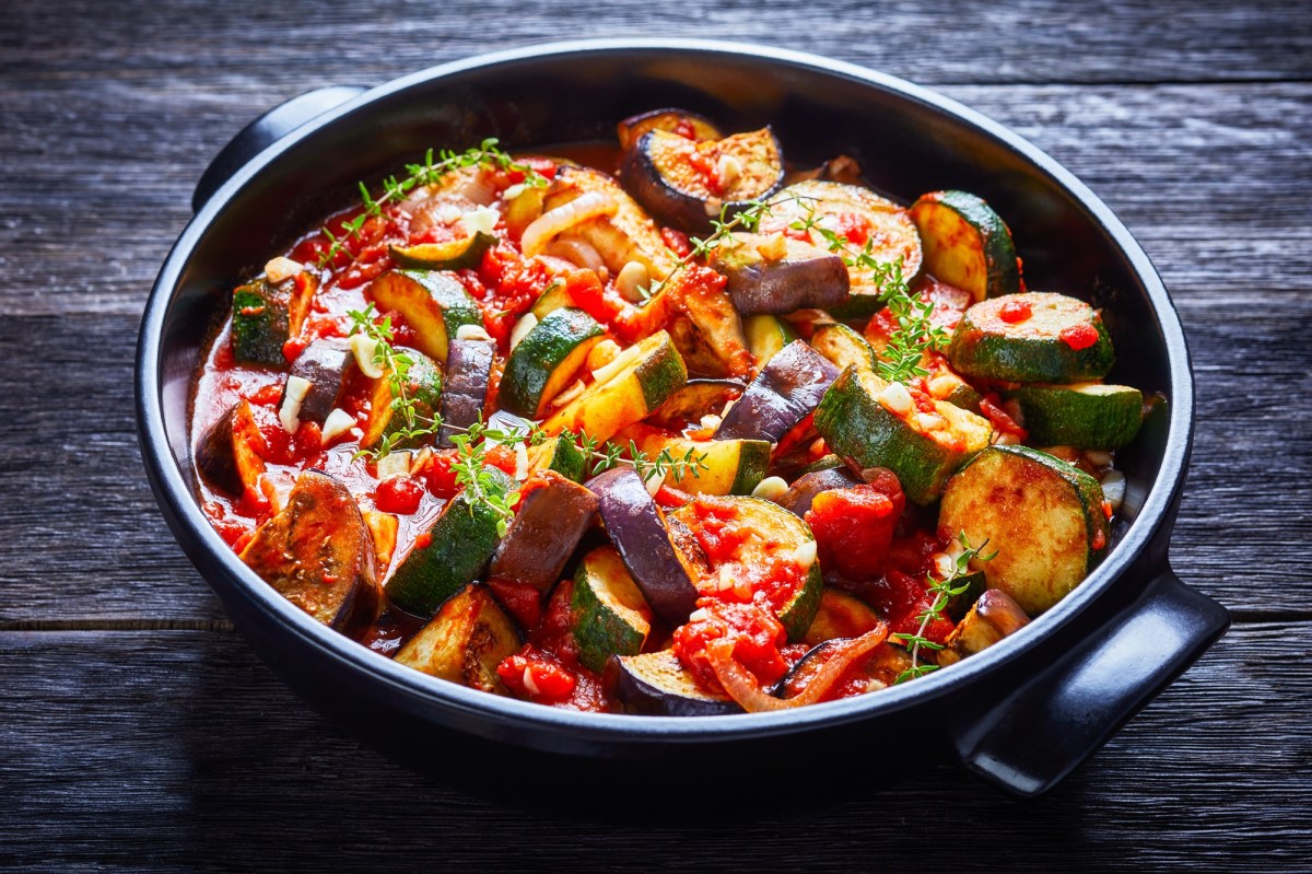 Ein GemÃ¼seeintopf mit Auberginen, Zucchini, Tomaten, Knoblauch und Zwiebeln, serviert in einer schwarzen Keramikschale und garniert mit KrÃ¤utern. Die Schale steht auf einem dunklen Holzuntergrund