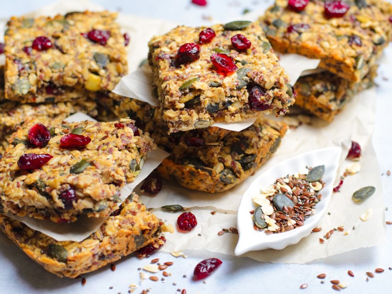 Einige Müsliriegel mit Cranberrys, Kürbiskernen und Kokos, die auf einer weißen Arbeitsfläche in kleinen Stapeln angerichtet sind. Es liegen getrocknete Cranberrys, Kürbiskerne und Leinsamen verstreut.