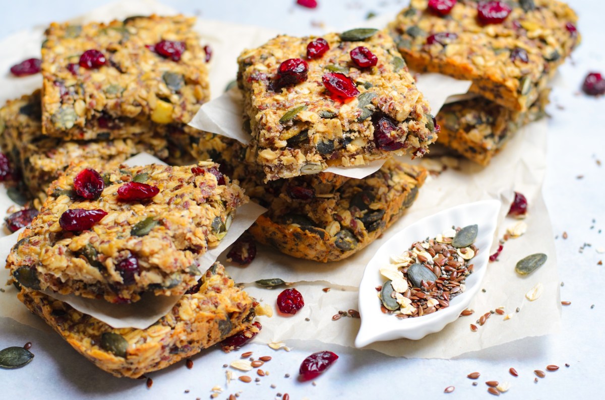 Einige MÃ¼sliriegel mit Cranberrys, KÃ¼rbiskernen und Kokos, die auf einer weiÃŸen ArbeitsflÃ¤che in kleinen Stapeln angerichtet sind. Es liegen getrocknete Cranberrys, KÃ¼rbiskerne und Leinsamen verstreut.