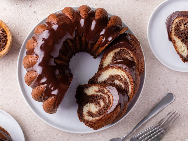 Draufsicht auf einen aufgeschnittenen Nutella-Marmorkuchen in Gugelhupfform. Der Kuchen steht auf einem weißen Teller. Rechts daneben steht ein weiterer Teller mit einem Stück Marmorkuchen mit Nutella. Links daneben steht eine Schale mit Schokosplittern und es liegen einige Kuchengabeln auf der Arbeitsfläche.