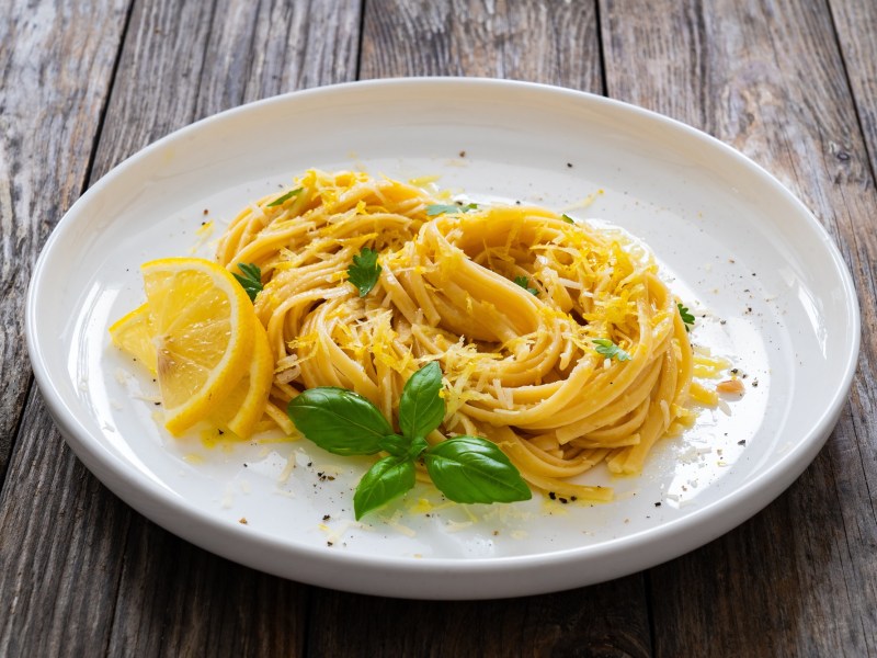 Eine Portion Pasta mit Zitrone und brauner Butter auf einem weißen, großen Teller, der auf einem Holztisch steht. Dekoriert ist die Pasta mit Zitrone und brauner Butter mit Zitronenscheiben, frischem Basilikum und geriebenem Parmesan.