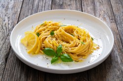 Eine Portion Pasta mit Zitrone und brauner Butter auf einem weißen, großen Teller, der auf einem Holztisch steht. Dekoriert ist die Pasta mit Zitrone und brauner Butter mit Zitronenscheiben, frischem Basilikum und geriebenem Parmesan.