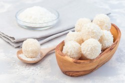 Eine Holzschale mit Raffaello-Energy-Balls auf hellem Untergrund. Im Hintergrund ist eine Glasschale mit Kokosraspel zu sehen, die auf einem hellgrauen Tuch steht. Neben der Holzschale liegt ein Holzlöffel mit Kokosraspel und einem einzelnen Raffaello-Energy-Ball.