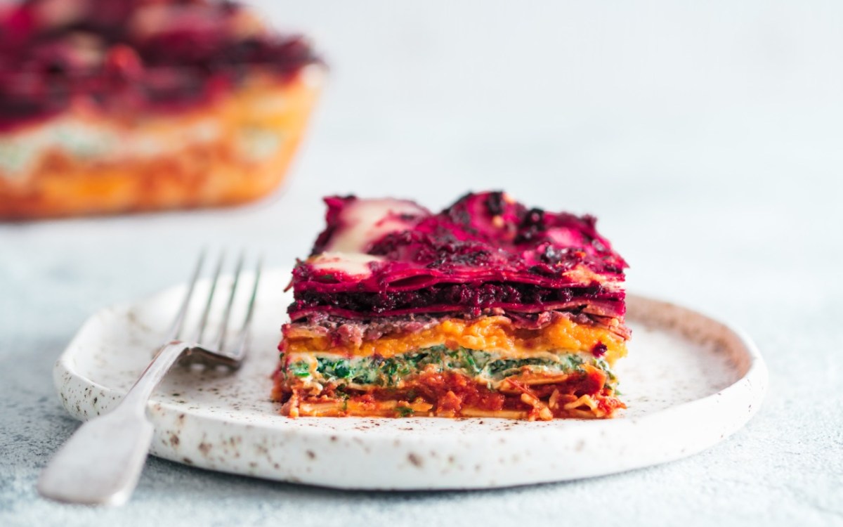 Ein Stück vegane Regenbogen-Lasagne auf einem Teller. Der Rest ist im Hintergrund zu sehen.