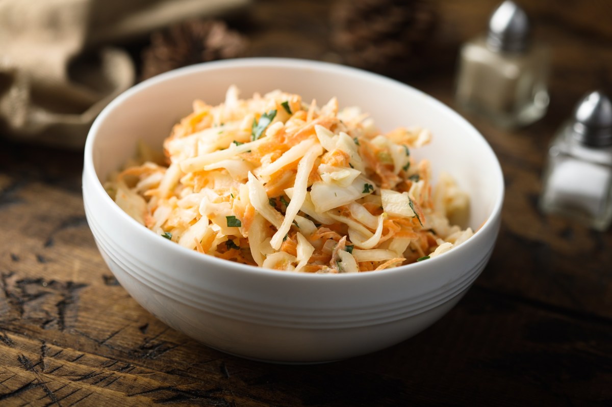 Eine SchÃ¼ssel, die Rettichsalat mit MÃ¶hren und Apfel enthÃ¤lt. Im Hintergrund sind verschwommen ein Salz- und Pfefferstreuer zu erkennen.