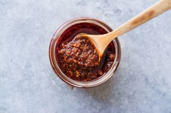 Draufsicht auf ein Glas Salsa Macha mit einem Holzlöffel darin. Das Glas mit der Salsa Macha steht auf einem hellgrauen Untergrund.