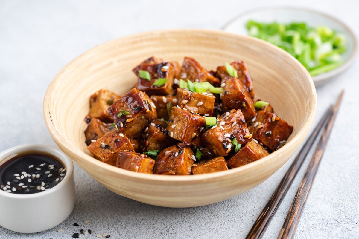 Teriyaki Tofu aus dem Airfryer in WÃ¼rfel geschnitten in einer kleinen Bambusschale. Sie sind mit FrÃ¼hlingszwiebeln und hellem sowie schwarzem Sesam angerichtet. Im Hintergrund steht ein kleiner Teller mit geschnittenen FrÃ¼hlingszwiebeln. Im Vordergrund steht eine Schale mit SojasoÃŸe.