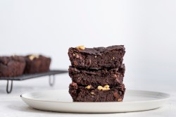 Drei Stücke vegane Kichererbsen-Brownies auf einem weißen großen Teller aufeinander gestapelt. Auf einem Gitter im Hintergrund sind weitere vegane Kichererbsen-Browniestücke zu sehen.