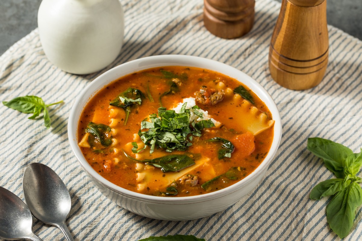 Lasagnesuppe in einer weiÃŸen SchÃ¼ssel steht auf einer gesteiften Tischdecke. Drumherum liegen ein LÃ¶ffel, BasilikumblÃ¤tter sowie ein Pfeffer- und Salzstreuer. Die Suppe ist ebenfalls mit Basilikum und KÃ¤se garniert.