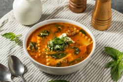 Lasagnesuppe in einer weißen Schüssel steht auf einer gesteiften Tischdecke. Drumherum liegen ein Löffel, Basilikumblätter sowie ein Pfeffer- und Salzstreuer. Die Suppe ist ebenfalls mit Basilikum und Käse garniert.