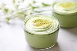 Ein kleines Dessertglas mit veganer Matcha-Weiße-Schokolade-Mousse getoppt mit etwas Matcha-Pulver. In Hintergrund sind verschwommen ein weiteres befülltes Dessertglas sowie einige Pflanzenstiele erkennbar.