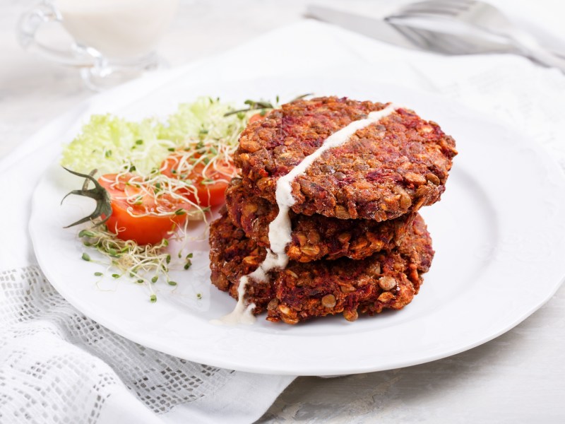 Vegane Rotkohlbratlinge mit Haferflocken liegen auf einem weißen Teller. Daneben liegt etwas Gartenkresse und eine halbierte Tomate.