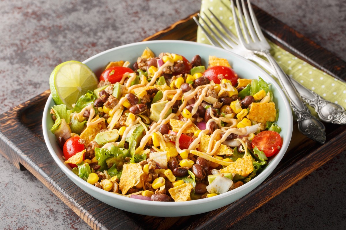 Bunter und veganer Nacho-Salat in einer weiÃŸen SchÃ¼ssel auf einem dunklen Holzbrett. Der Salat besteht aus Nachos, Kidneybohnen, Nachos, frischem Salat, Mais und Tomaten. Links neben der Schale liegen zwei Gabeln auf einer Serviette.