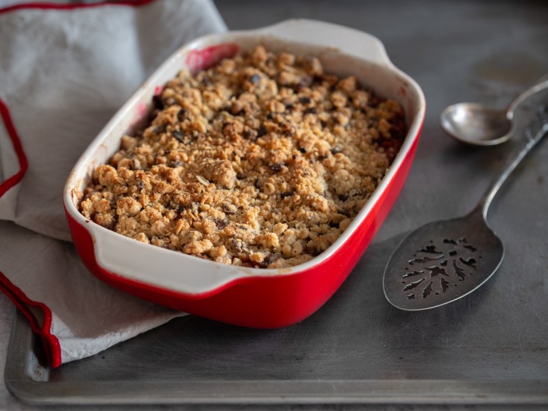 Veganer Porridgeauflauf in einer weiÃŸ-roten Auflaufform. Man sieht am Rand, dass er mit Beeren zubereitet ist. Links neben ihm liegt ein weiÃŸ-rotes Tuch, rechts zwei LÃ¶ffel.