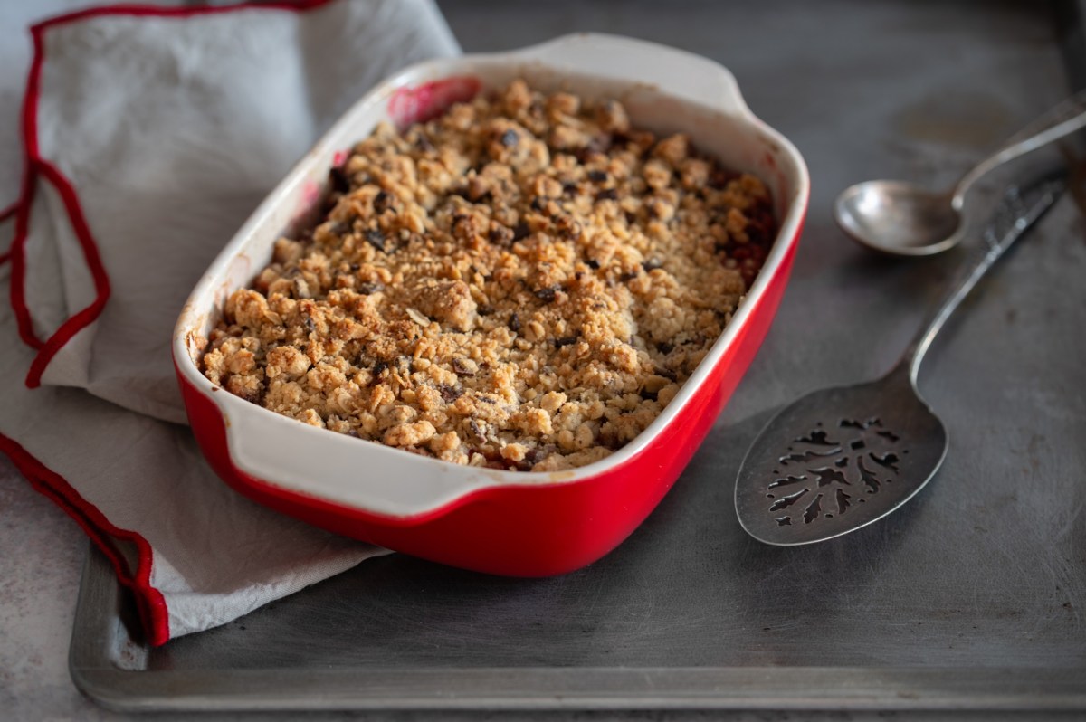 Veganer Porridgeauflauf in einer weiÃŸ-roten Auflaufform. Man sieht am Rand, dass er mit Beeren zubereitet ist. Links neben ihm liegt ein weiÃŸ-rotes Tuch, rechts zwei LÃ¶ffel.