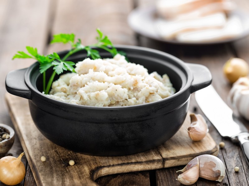 Ein schwarzes Töpfchen mit veganem Schmalz, garniert mit frischer Petersilie, angerichtet auf einem Holzbrett. Daneben liegt ein Messer, sowie Knoblauchzehen und Pfefferkörner.