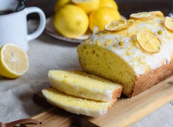 Ein angeschnittener Zitronen-Kastenkuchen mit Puderzucker und Zitronenscheiben dekoriert. Im Hintergrund ein Teller Zitronen und eine Tasse.