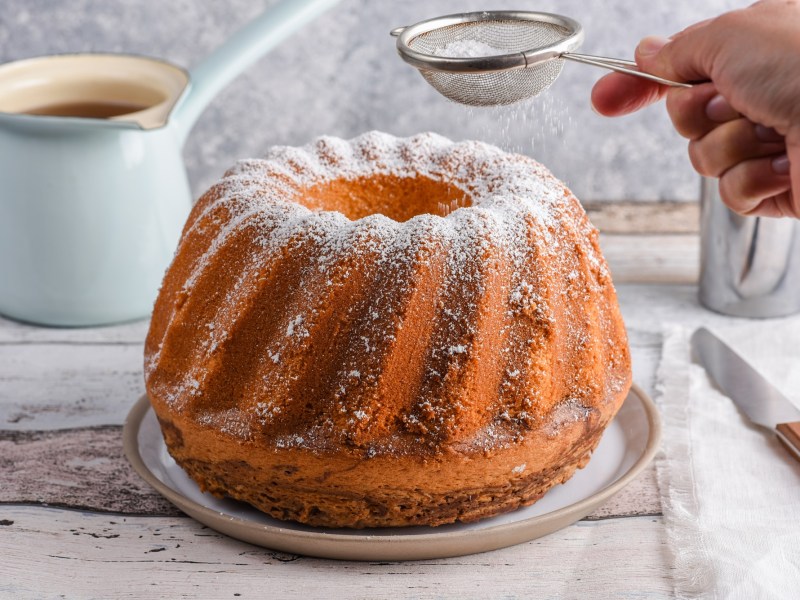 Ein Amaretto-Kuchen in Gugelhupfform, der auf einem Teller steht und gerade mit Puderzucker bestäubt wird. Neben dem Kuchen liegt auf einem Stück Stoff ein Messer. Im Hintergrund steht ein gefülltes Gefäß.