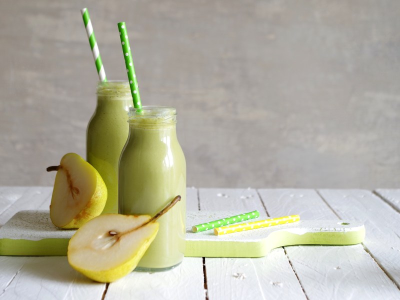 Zwei kleine Glasflaschen mit hellgrünem Birnen-Feldsalat-Smoothie, die auf einem weißen Holzbrett stehen. Auf dem Brett liegen außerdem ein gelber und ein grüner Strohhalm und es liegen 2 Birnenhälften mit im Bild. In den Glasflaschen steckt auch jeweils ein grüner Strohhalm.