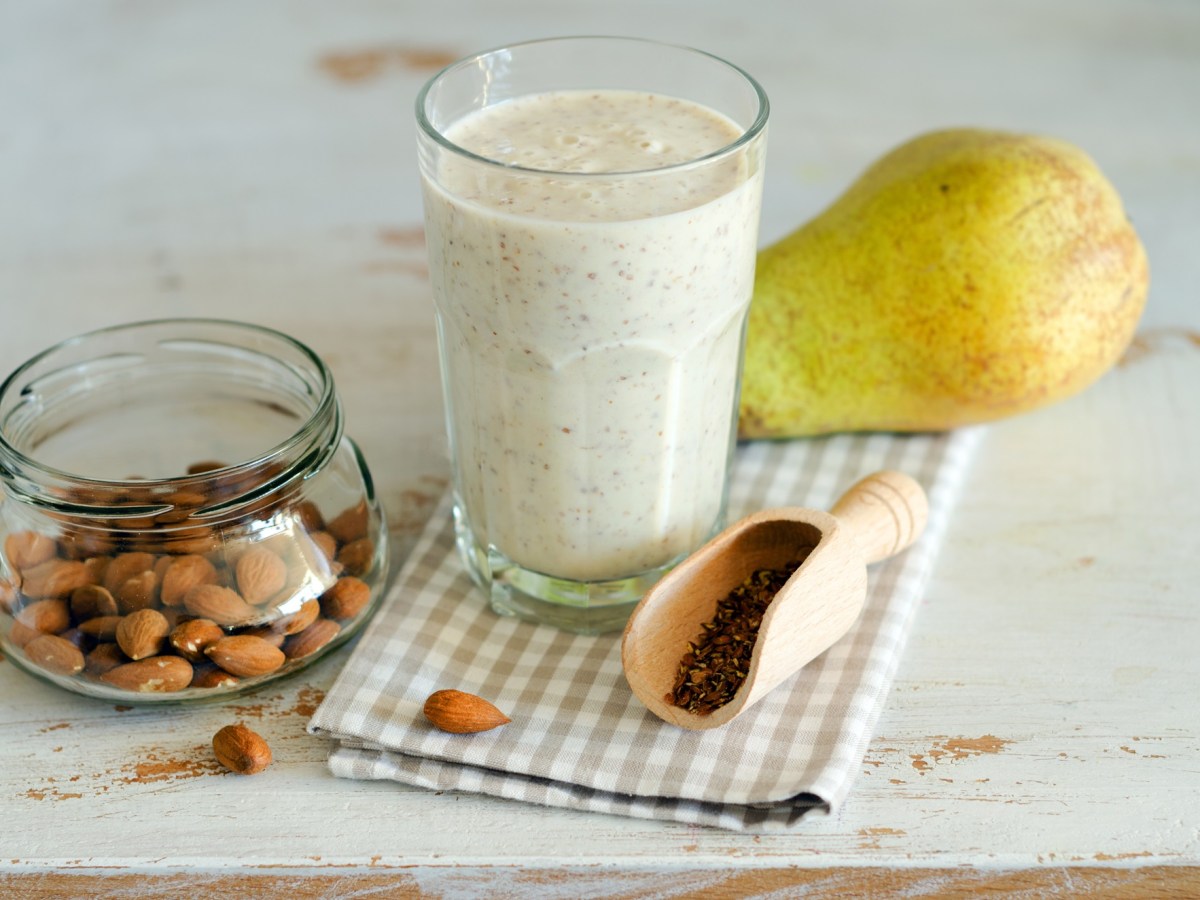 Ein Glas mit einem cremigen Birnen-Mandel-Smoothie. Das Klas steht auf einem karierten Küchentuch. Außerdem liegt eine Birne im Bild. Neben dem Glas steht eine Glasschale mit Mandeln. Zusätzlich ist eine kleine Holzschaufel mit ein paar Leinsamen zu sehen.