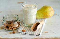 Ein Glas mit einem cremigen Birnen-Mandel-Smoothie. Das Klas steht auf einem karierten Küchentuch. Außerdem liegt eine Birne im Bild. Neben dem Glas steht eine Glasschale mit Mandeln. Zusätzlich ist eine kleine Holzschaufel mit ein paar Leinsamen zu sehen.