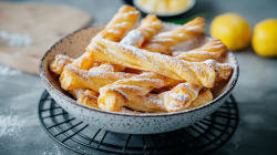 Eine Schüssel mit Blätterteig-Quarkstangen, die mit Puderzucker bestäubt sind. Die Schüssel steht auf einem Gitter-Untersetzer. Im Hintergrund liegt ein Holzbrett, auf dem Mehl oder Puderzucker verstreut ist. Außerdem liegen im Hintergrund zwei Zitronen.