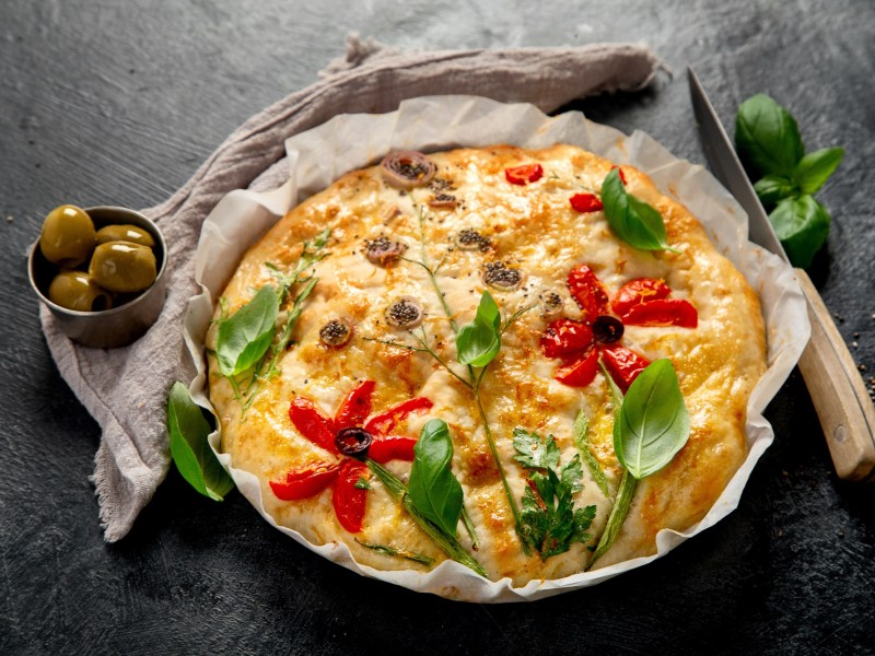 Ein dekoriertes Blumen-Focaccia liegt auf einem runden Teller auf Backpapier. Es ist mit Basilikum, Tomaten und Oliven. LInks neben dem Focaccia liegt eine kleine Schale mit Oliven, rechts ein scharfes Messer.