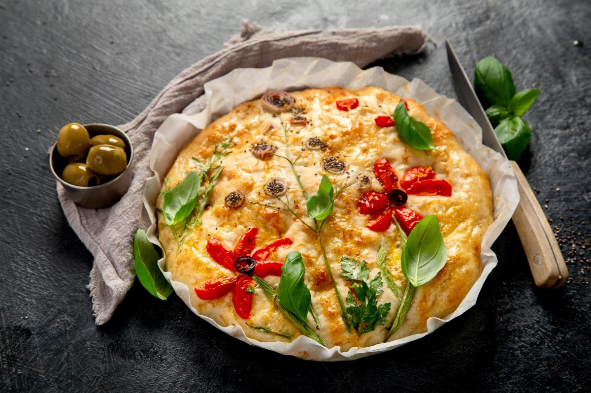 Ein dekoriertes Blumen-Focaccia liegt auf einem runden Teller auf Backpapier. Es ist mit Basilikum, Tomaten und Oliven. LInks neben dem Focaccia liegt eine kleine Schale mit Oliven, rechts ein scharfes Messer.