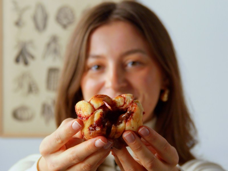 Unsere Köchin hält einen auseinandergezogenen Brioche-Muffin in die Kamera. Man sieht deutlich die Schokoladenfüllung. Im Hintergrund hängt ein Bild mit verschiedenen Wurzelgewächsen.